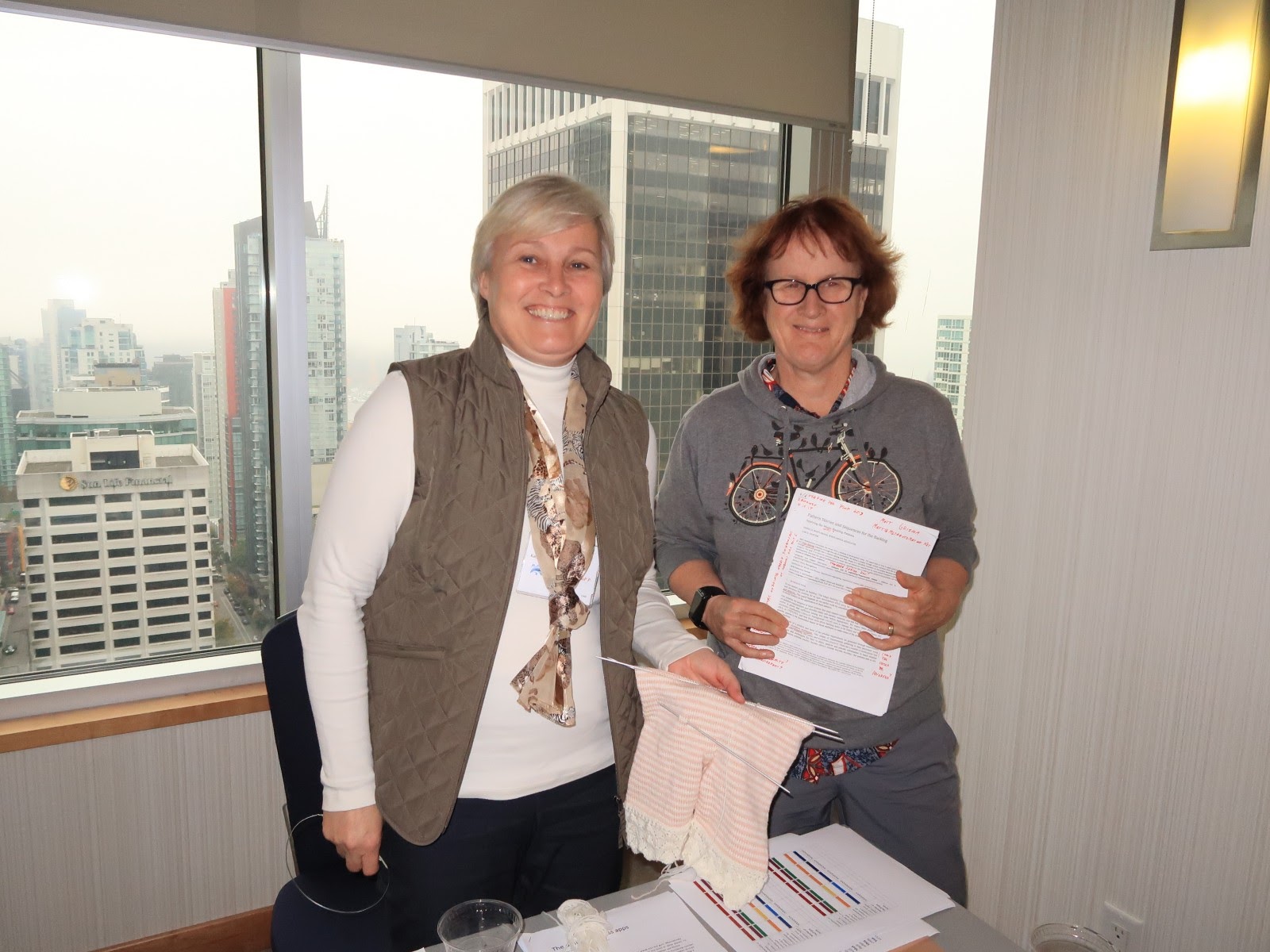 The two authors standing and smiling in front of a window onto the city skyline. Lise, on the left, holds some knitting; Rebecca a marked up manuscript.