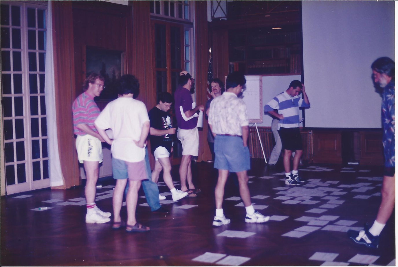 A group of men wearing shorts and casual shirts in the library at Allerton. Rectangular papers are strewn on the floor. Some are looking at clusters of papers. Some are talking to each other. One man is looking at a grouping of papers and scratching his head.