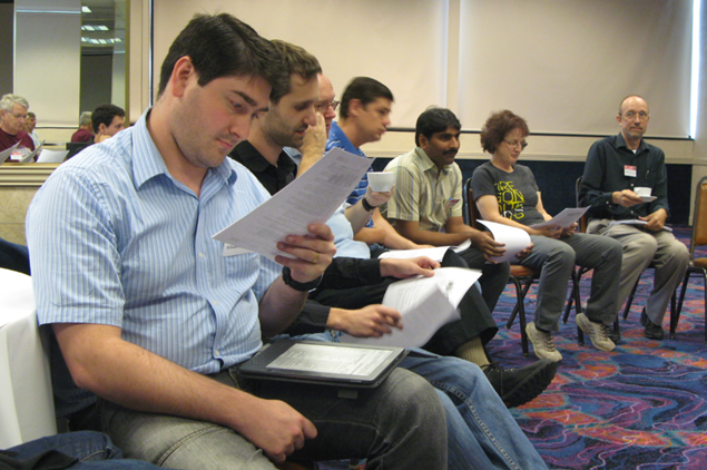 People seated in chairs in a writers' workshop intently looking at manuscripts during a discussion. A young Eduardo Guerra wearing a blue striped short sleeved shirt and jeans and holding a manuscript, is closest to the camera. In the chair second to the right is Rebecca holding a manuscript and wearing a tshirt and jeans.